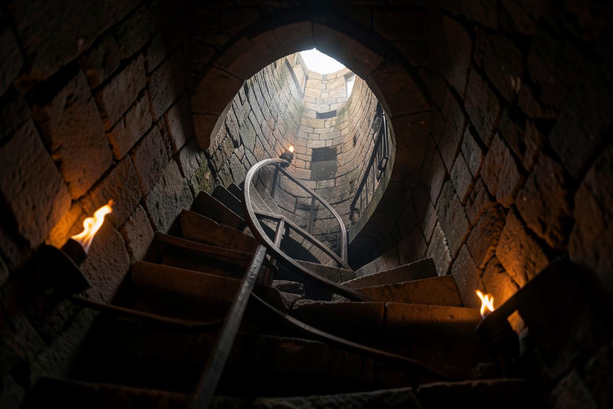 Escada em caracol de pedra numa torre antiga, iluminada por tochas, subindo em direção à luz