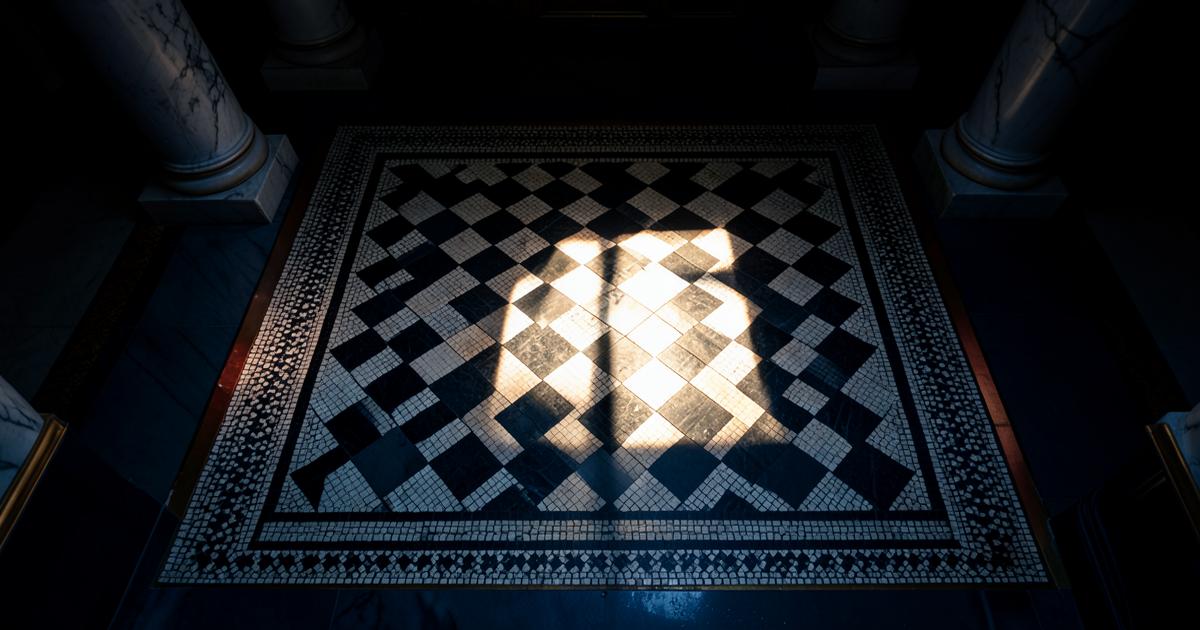Pavimento mosaico maçônico de quadrados pretos e brancos com orla entalhada, iluminado por um feixe de luz no centro do templo